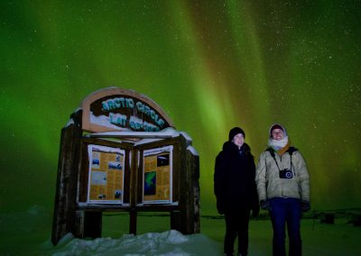 two people standing beside an arctic circle sign, aurora borealis in the night sky
