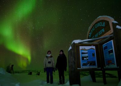 two people standing beside an arctic circle sign, aurora borealis in the night sky